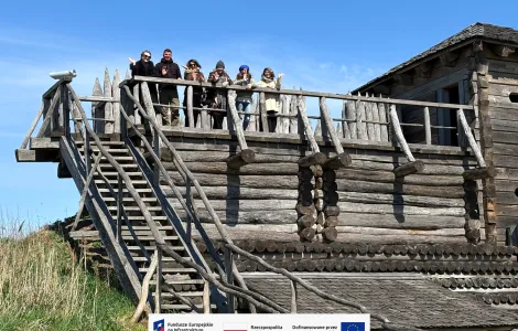 Sześć osób stoi na rekonstrukcji budowli z drewnianych bali i macha w stronę aparatu. Po prawej stronie widać fragment drewnianej wieży. W tle rozciąga się jasne, bezchmurne niebo, a wokół rośnie trawa. Na dole widoczne logotypy Rzeczypospolitej Polskiej oraz Funduszy Europejskich.