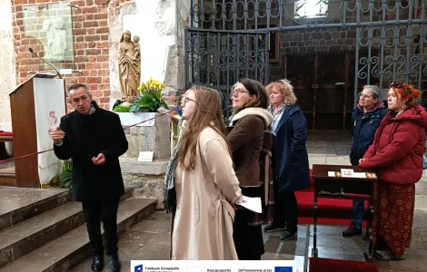 Grupa kilku osób stoi we wnętrzu zabytkowego kościoła z cegły i kamienia. Mężczyzna ubrany na czarno gestykuluje, a pozostali słuchają i patrzą w jego stronę. W tle widać ozdobną metalową bramę, rzeźbę religijną z kwiatami oraz ambonę, a na podłodze leżą czerwone dywany. Na dole widoczne logotypy Rzeczypospolitej Polskiej oraz Funduszy Europejskich.
