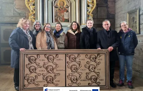 Grupa osób stoi we wnętrzu drewnianego kościoła przed ozdobionym ołtarzem z ikoną świętego. Wszyscy ustawieni są w jednym rzędzie i trzymają duży obraz na desce przedstawiający rzędy czaszek ze skrzyżowanymi kośćmi oraz łacińskie napisy, min. „Memento mori”. W tle widoczne są bogato zdobione elementy ołtarza oraz drewniane ściany świątyni. Na dole widoczne logotypy Rzeczypospolitej Polskiej oraz Funduszy Europejskich.