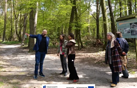 Grupa kilku osób stoi na leśnej ścieżce wśród wysokich drzew. Mężczyzna w niebieskiej marynarce wskazuje ręką w bok, a pozostali uczestnicy spaceru słuchają i patrzą w jego kierunku. W tle widoczna jest tablica informacyjna oraz słoneczna, zielona sceneria lasu. Na dole widoczne logotypy Rzeczypospolitej Polskiej oraz Funduszy Europejskich.