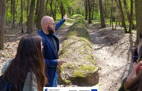 Grupa osób stoi na leśnej ścieżce obok długiego grobowca megalitycznego. Mężczyzna na pierwszym planie gestem ręki wskazuje kierunek wzdłuż nasypu, podczas gdy pozostali słuchają. Wokół rosną wysokie drzewa, a słoneczne światło przebija się przez wiosenne liście. Na dole widoczne logotypy Rzeczypospolitej Polskiej oraz Funduszy Europejskich.