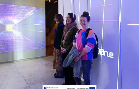 Trzy osoby stoją przy ścianie w nowoczesnej, multimedialnej przestrzeni wystawowej. Na ścianie obok nich widoczna jest projekcja świetlna z geometrycznym wzorem, a pionowe linie świetlne tworzą futurystyczny klimat. Dwie osoby uśmiechają się w stronę aparatu. Na dole widoczne logotypy Rzeczypospolitej Polskiej oraz Funduszy Europejskich.