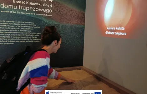 Osoba w muzeum pochyla się nad interaktywną ekspozycją z piaskiem, dotykając jej dłonią. Na ścianie obok wyświetlany jest obraz glinianej amfory z podpisem „Amfora kulista / Globular amphora”. W tle widoczna jest tablica informacyjna dotycząca stanowiska archeologicznego w Brześciu Kujawskim. Na dole widoczne logotypy Rzeczypospolitej Polskiej oraz Funduszy Europejskich.