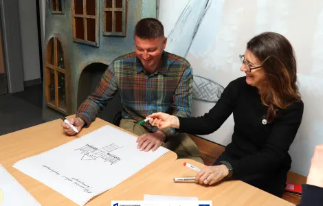 Dwie osoby siedzą przy stole i wspólnie zapisują propozycje na dużej kartce papieru. Mężczyzna w koszuli w kratę trzyma czarny marker i uśmiecha się, patrząc na szkic budynku na kartce. Kobieta obok niego, ubrana w czarny sweter i okulary, podaje mu zielony marker, również uśmiechnięta. Na stole leżą dodatkowe markery i kartki. W tle widać dekoracyjną ścianę z elementami przypominającymi fasadę domu. Na dole widoczne logotypy Rzeczypospolitej Polskiej oraz Funduszy Europejskich.