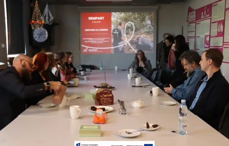 Grupa kilkunastu osób siedzi przy długim stole w sali konferencyjnej podczas spotkania. Na końcu stołu wyświetlany jest slajd prezentacji z tytułem „REMPART - Heritage as a Bridge”, a jedna z uczestniczek prowadzi dyskusję. Na stole stoją filiżanki, talerzyki z ciastkami i napojami. Uczestnicy słuchają i piją kawę, a na ścianach widać plansze informacyjne. Na dole widoczne logotypy Rzeczypospolitej Polskiej oraz Funduszy Europejskich.
