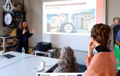 Kobieta prowadzi prezentację przed kilkoma słuchaczami w sali konferencyjnej. Stoi przy ekranie projekcyjnym i gestykuluje rękami, wyjaśniając temat. Na slajdzie widnieje tytuł „What volunteering can accomplish” oraz zdjęcia pokazujące efekt „przed i po” renowacji budynku. Uczestnicy siedzą przy stole, na którym stoją kubki, talerzyki i butelka wody, i uważnie słuchają prezentacji. Na dole widoczne logotypy Rzeczypospolitej Polskiej oraz Funduszy Europejskich.