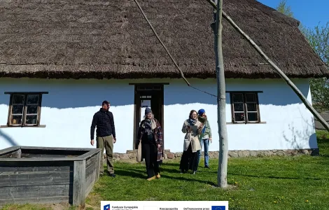 Cztery osoby stoją przed tradycyjną białą chatą z dachem krytym strzechą. Na pierwszym planie widać żuraw studzienny. Nad całą sceną rozciąga się bezchmurne, intensywnie niebieskie niebo, a wokół domu rośnie zielona trawa. Na dole widoczne logotypy Rzeczypospolitej Polskiej oraz Funduszy Europejskich.