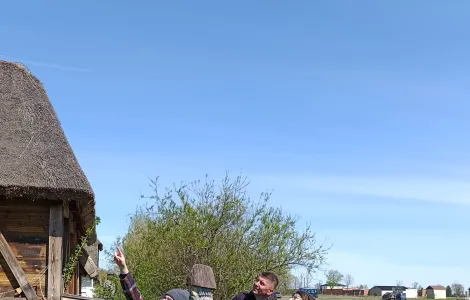 Cztery osoby stoją na trawie obok drewnianej chaty krytej strzechą. Jedna z nich wskazuje ręką w górę, a pozostali patrzą w tym samym kierunku. W tle widać krzewy, pole uprawne oraz kilka budynków, a nad całą sceną rozciąga się jasne, bezchmurne niebo. Na dole widoczne logotypy Rzeczypospolitej Polskiej oraz Funduszy Europejskich.