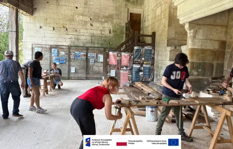 Grupa osób pracuje przy obróbce drewna w dużym, półotwartym pomieszczeniu o kamiennych ścianach. Na pierwszym planie trzy młode osoby stoją przy drewnianych stołach warsztatowych i ręcznie strugają oraz dopasowują elementy z drewna. W tle widać kolejne osoby przy pracy oraz tablice informacyjne ustawione pod ścianą. Po prawej stronie znajduje się duży, kamienny kominek. Na dole obrazu widoczny jest pasek z logotypami Funduszy Europejskich, Rzeczypospolitej Polskiej i Unii Europejskiej.