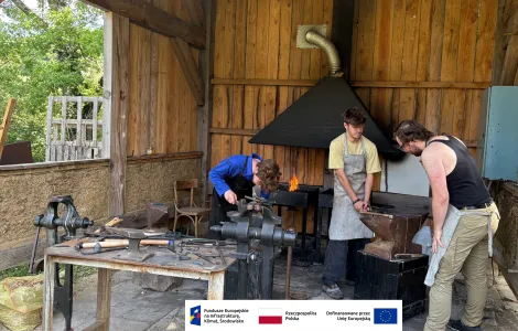Trzy osoby pracują w kuźni w drewnianej, półotwartej szopie. Na środku pali się ogień w palenisku pod metalowym okapem. Jedna osoba pochyla się nad imadłem, druga trzyma rozgrzany metal przy kowadle, a trzecia obserwuje. Wokół znajdują się narzędzia kowalskie, takie jak młotki, szczypce i kowadło ustawione na solidnej podstawie. Na dole obrazu widoczny jest pasek z logotypami Funduszy Europejskich, Rzeczypospolitej Polskiej i Unii Europejskiej.