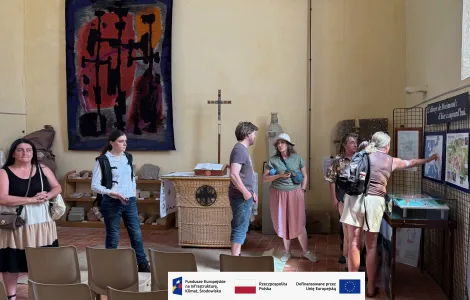 Grupa osób zwiedza wnętrze jasnego budynku. Kilka osób stoi przy tablicach informacyjnych i ogląda mapy oraz plansze wystawowe, jedna osoba wskazuje na jeden z elementów ekspozycji. W tle widoczny jest ołtarz z krzyżem, rzeźbą oraz tkaniną artystyczną zawieszoną na ścianie. W pomieszczeniu ustawione są także krzesła. Na dole obrazu znajduje się pasek z logotypami Funduszy Europejskich, Rzeczypospolitej Polskiej i Unii Europejskiej.