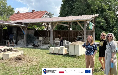Trzy kobiety stojące na trawie przed zadaszoną, drewnianą wiatą warsztatową. Pod wiatą dwie osoby pracują przy obróbce jasnych, kamiennych bloków, a obok leżą stosy elementów i narzędzia. W tle znajduje się dom z czerwonym dachem oraz drzewa. Na pierwszym planie ustawione są duże, jasne bloki. Na dole obrazu widoczny jest pasek z logotypami Funduszy Europejskich, Rzeczypospolitej Polskiej i Unii Europejskiej.